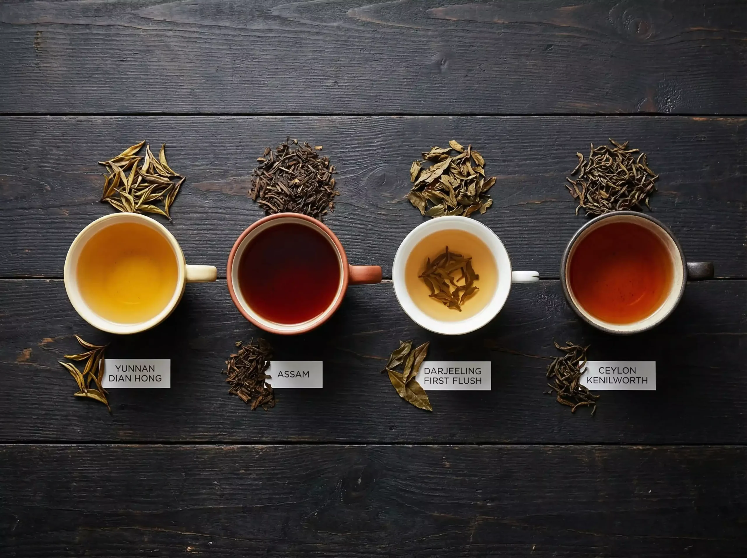 A dark wooden table showing four cups of black tea side by side: bright golden Yunnan Dian Hong, bold dark red Assam