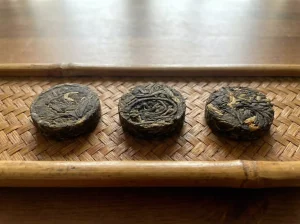 Close-up of several Steeped Roots 2g mini tea cakes, small round compressed discs including raw pu'er (greenish), ripe pu'er (dark brown), and Yunnan red tea (reddish), neatly arranged on a bamboo tray or wooden table