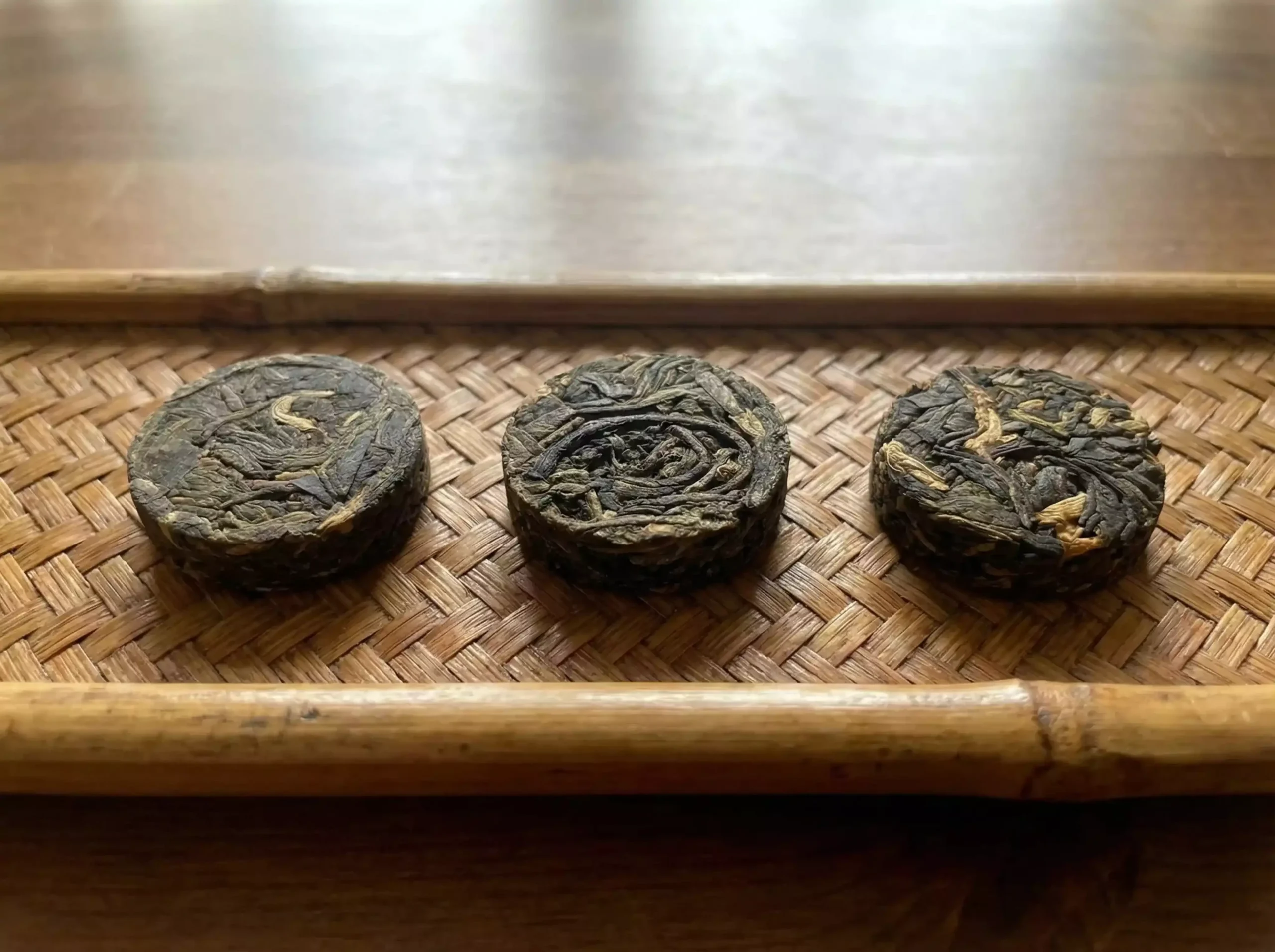 Close-up of several Steeped Roots 2g mini tea cakes, small round compressed discs including raw pu'er (greenish), ripe pu'er (dark brown), and Yunnan red tea (reddish), neatly arranged on a bamboo tray or wooden table