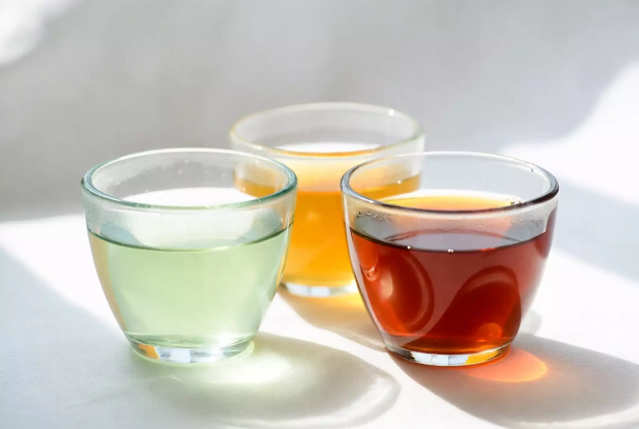 Side-by-side clear glass cups displaying the range of tea liquor colors: pale jade green, golden, amber, ruby red, and deep reddish-brown
