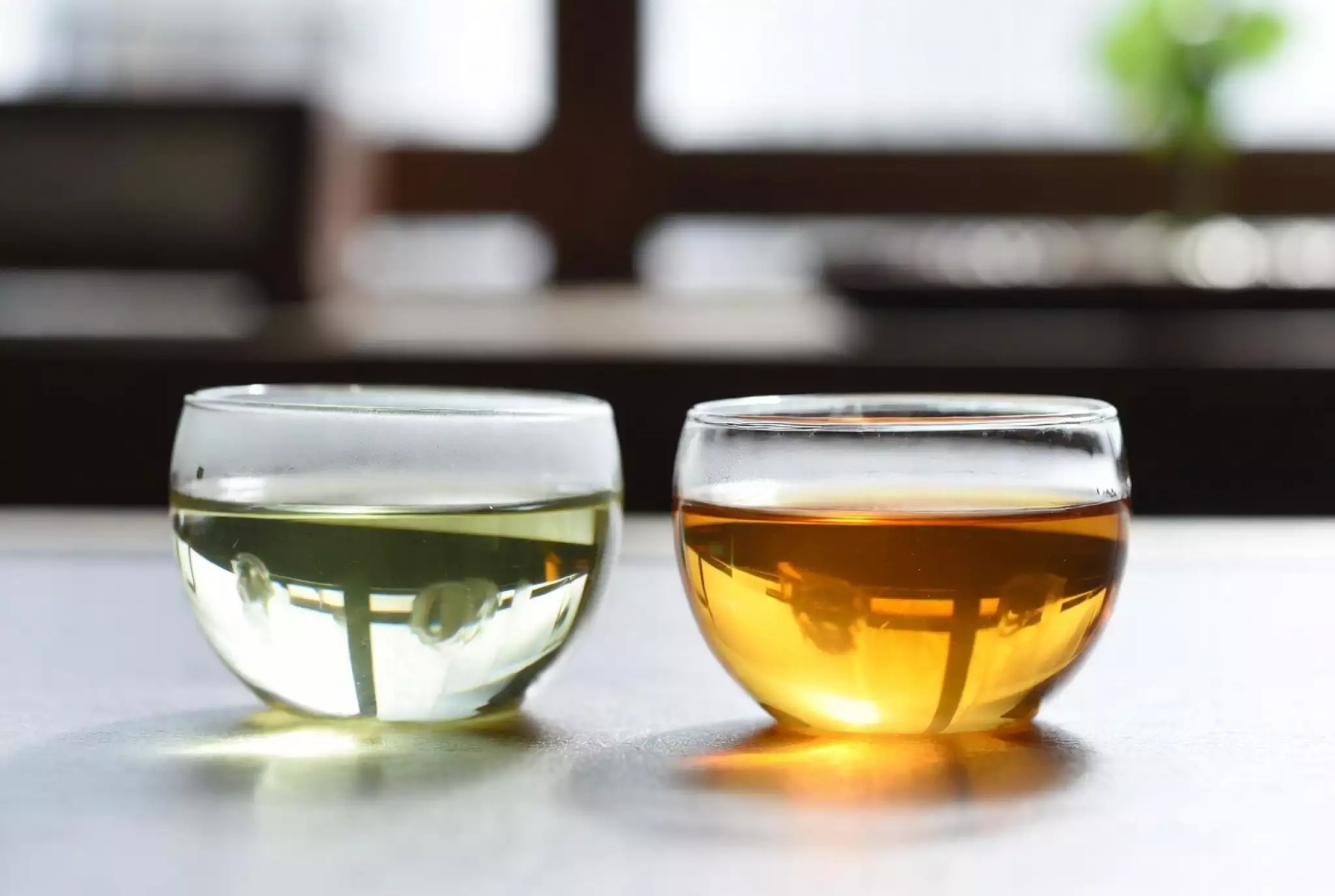 Two glass tea cups side by side in a minimalist Gongfu tea ceremony setting. Left cup: pale yellow-green bright tea liquor. Right cup: rich amber golden tea liquor.