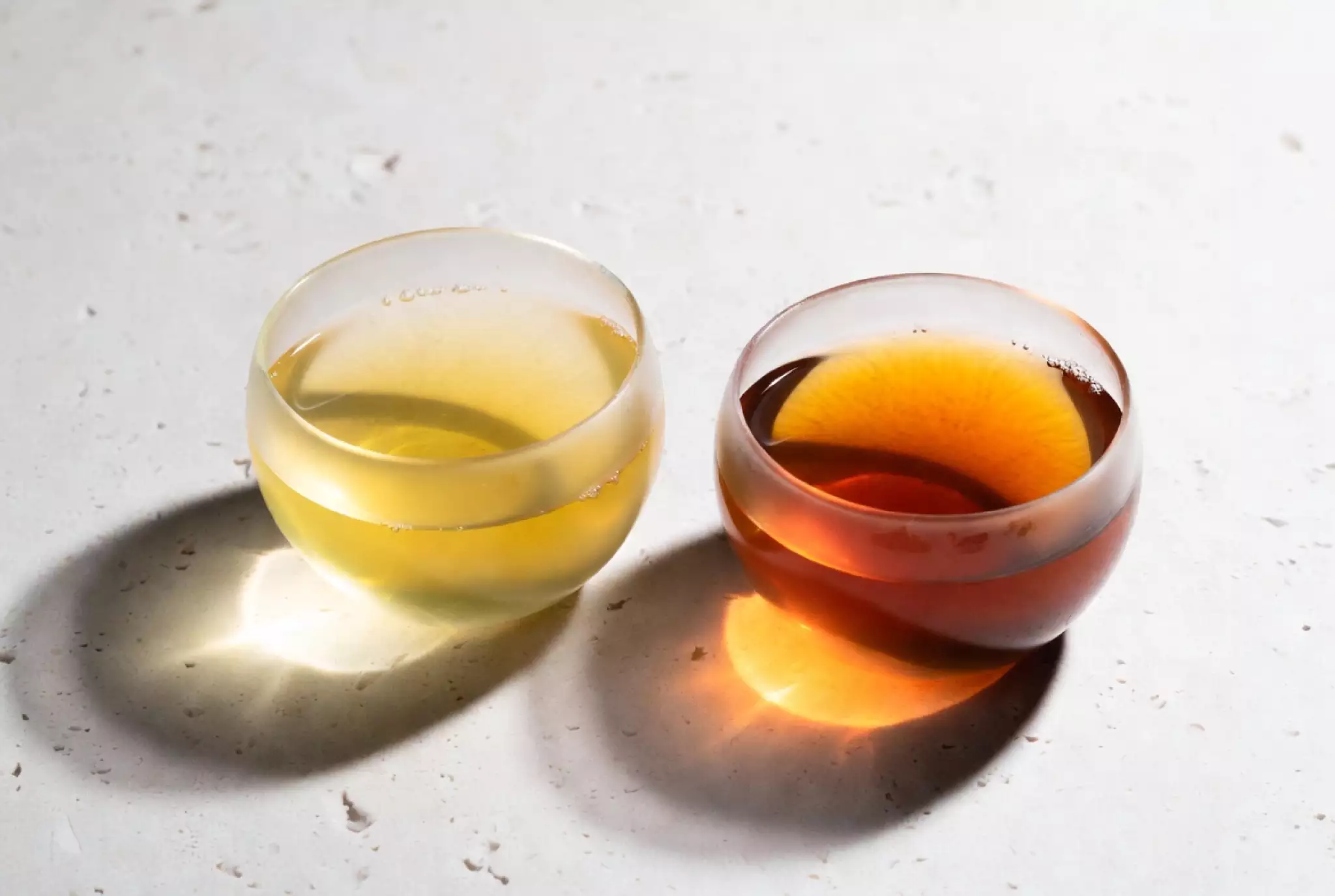 Two translucent glass tea cups side by side on a minimalist white stone surface. The left cup contains bright, pale-yellow young Puerh tea soup. The right cup contains deep amber, glowing aged Puerh tea soup