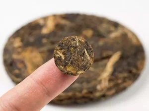 Close-up of a tiny 2g compressed tea disc on a fingertip, showing tea leaf texture and miniature scale.