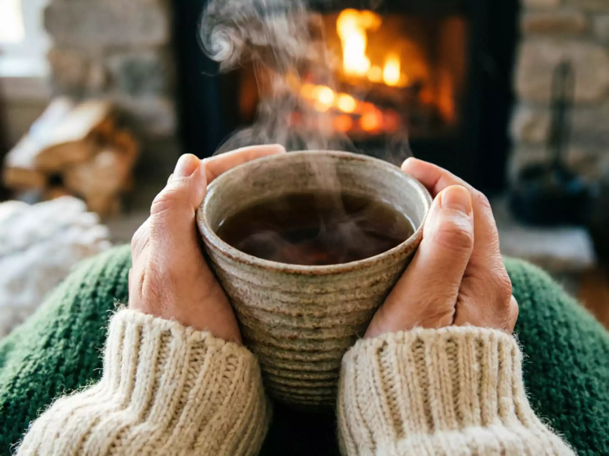 Close-up of hands in a wool sweater holding a warm ceramic mug of tea by a winter fireplace